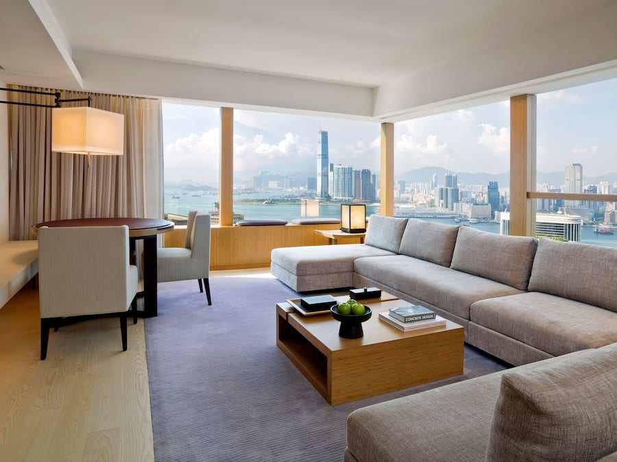 A modern hotel room with a large window offering a city view, a sofa, a coffee table, and a desk with a lamp.