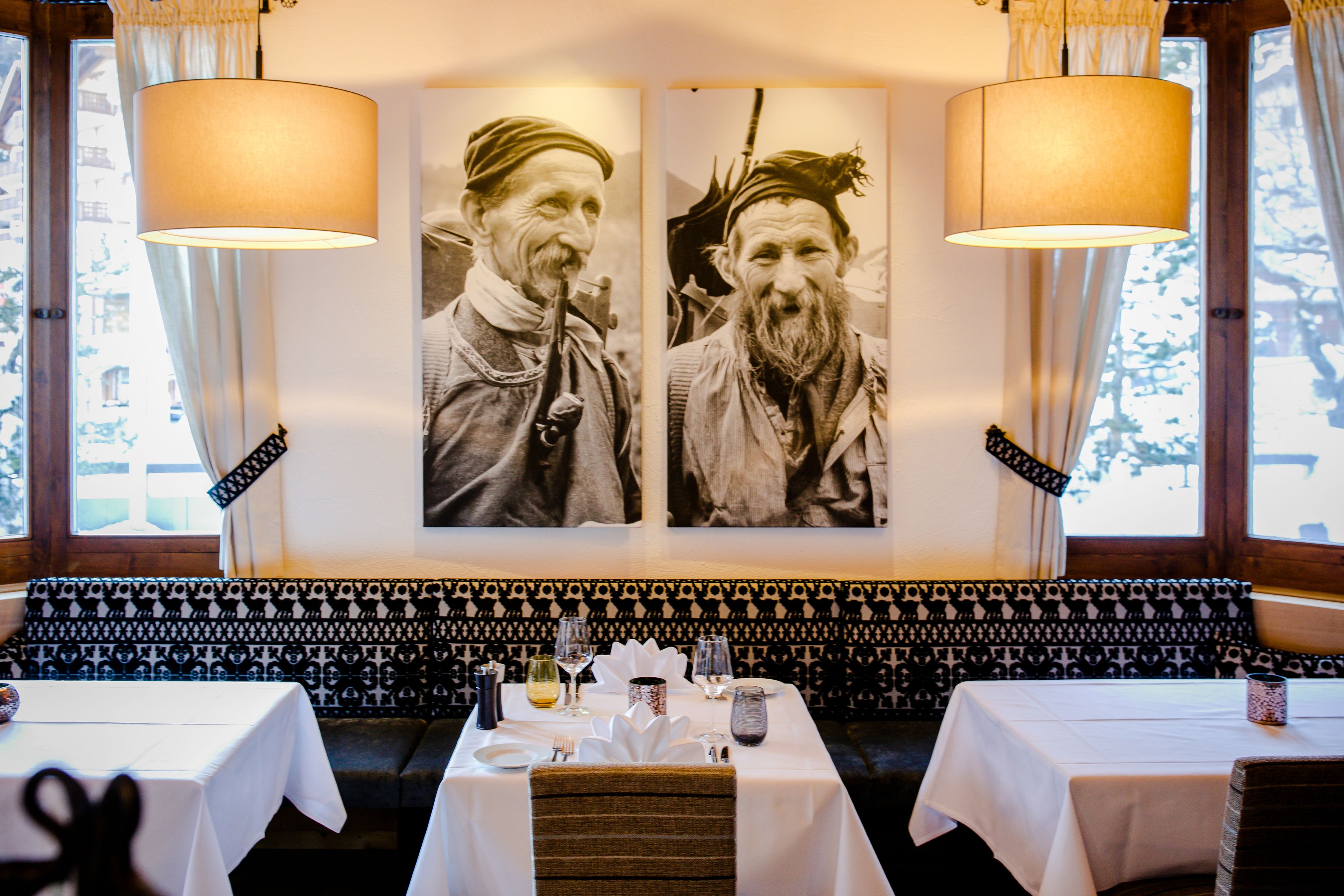 Un restaurante con un banco de asientos a cuadros blanco y negro, dos lámparas grandes fijadas a la pared y una mesa con mantel blanco, copas de vino y un jarrón.