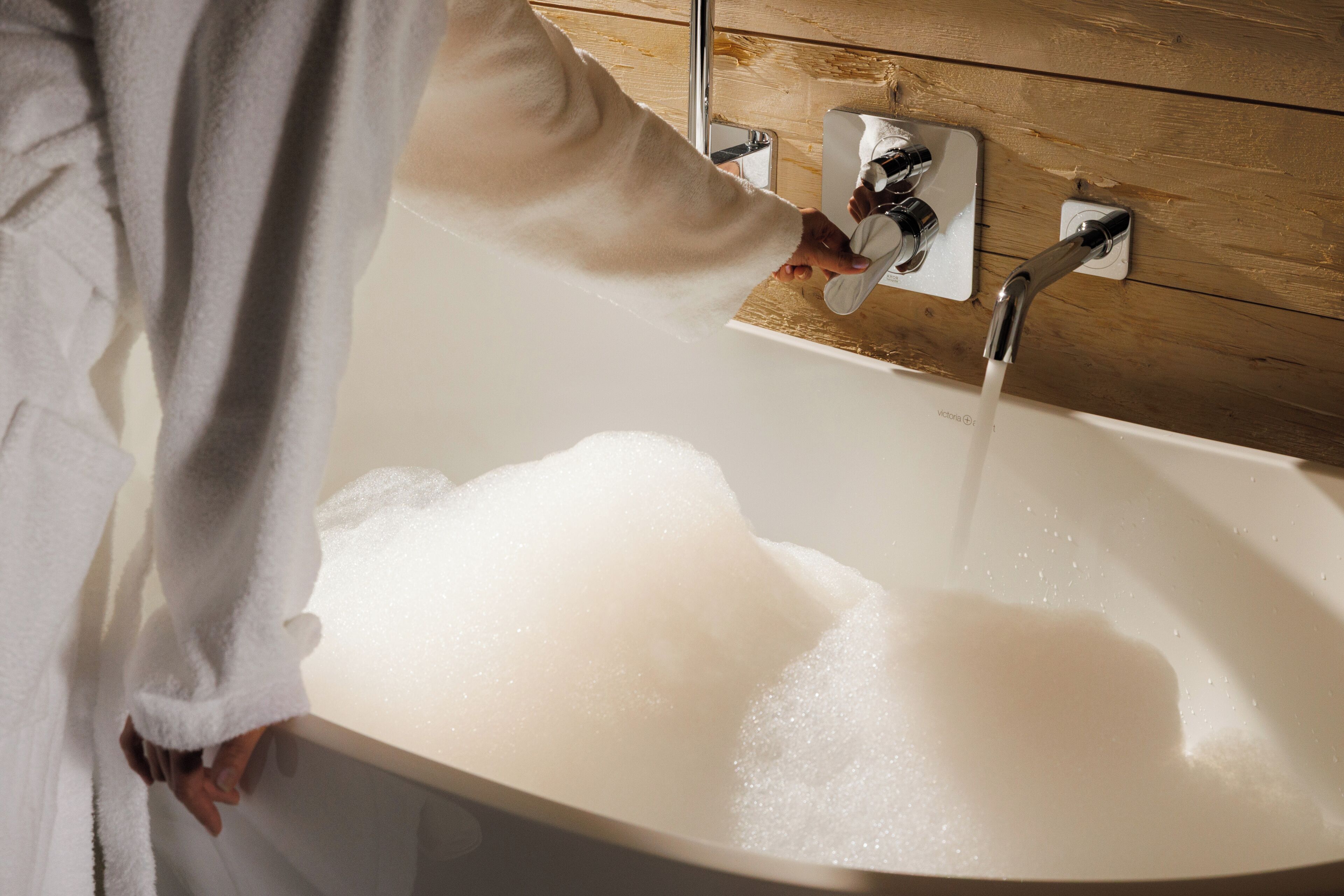 A person in a white robe standing by a bathtub filled with bubbles, with a wooden wall in the background.