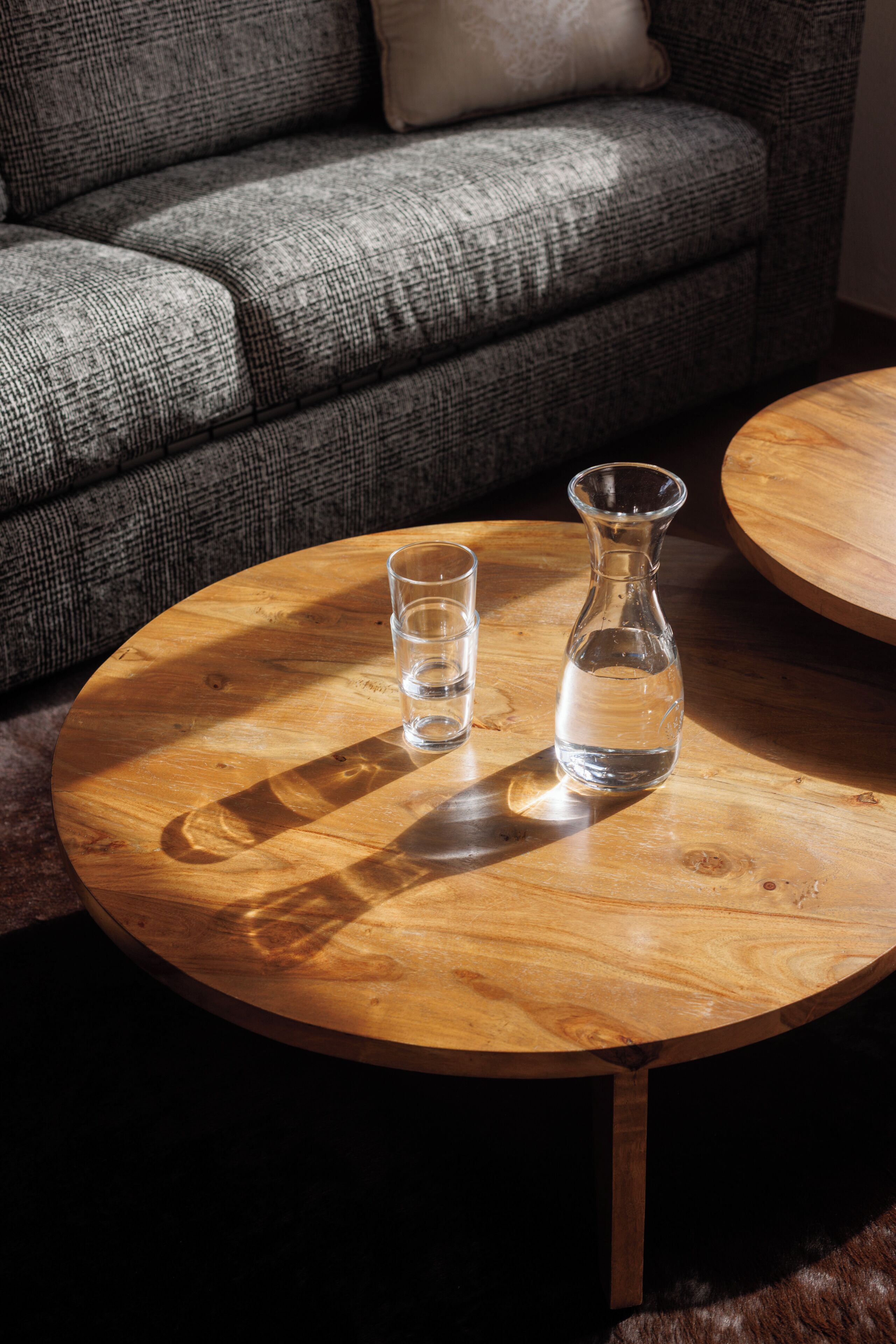 Una mesa redonda de madera con una jarra de vidrio transparente y un vaso encima, sobre un piso de madera.