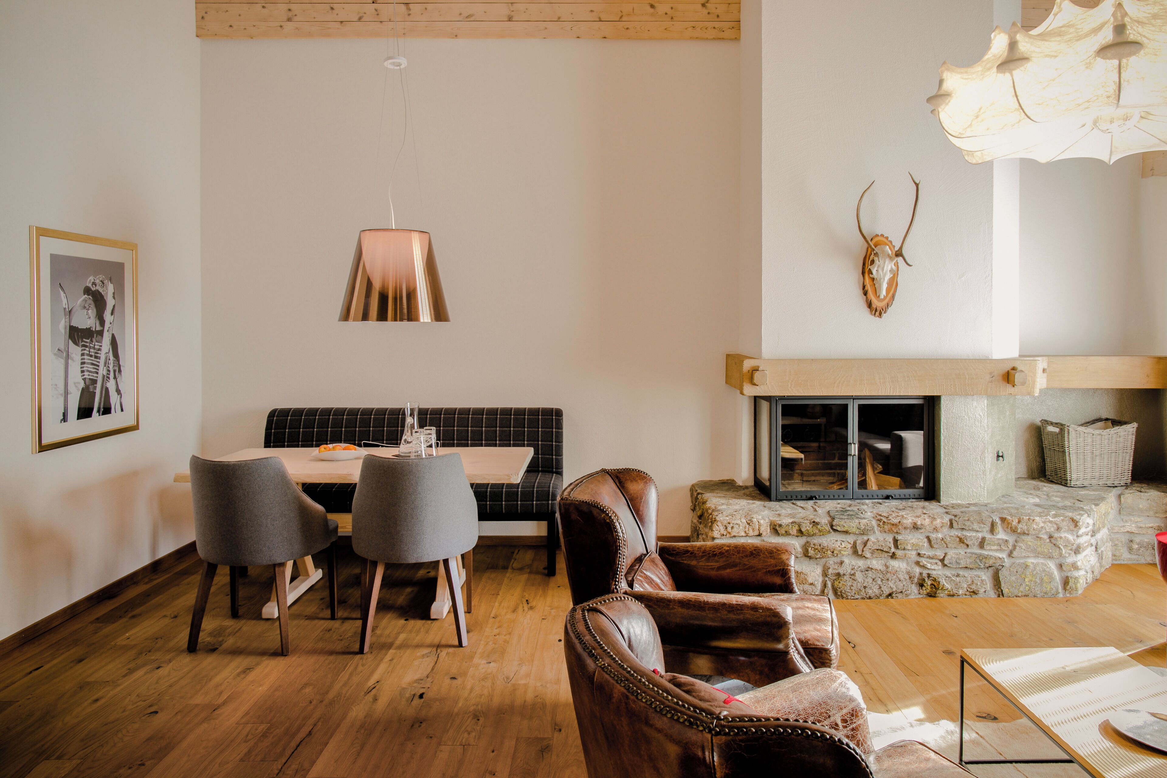 A dining area with a table, chairs, and a bench. There is a fireplace with a stone surround and a mounted deer skull.