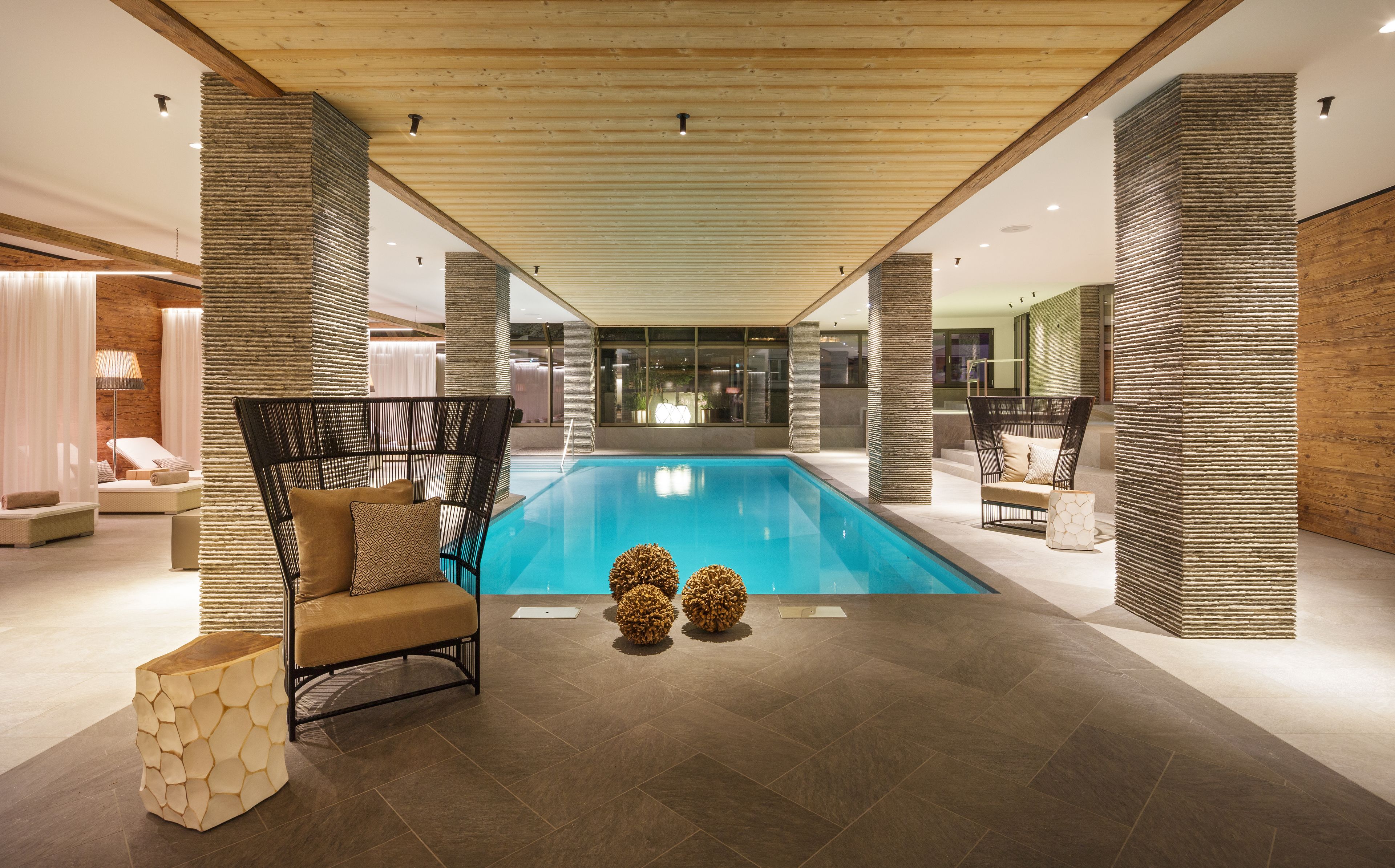 A modern indoor pool area with a wooden ceiling, lounge chairs, and a view of the outside.