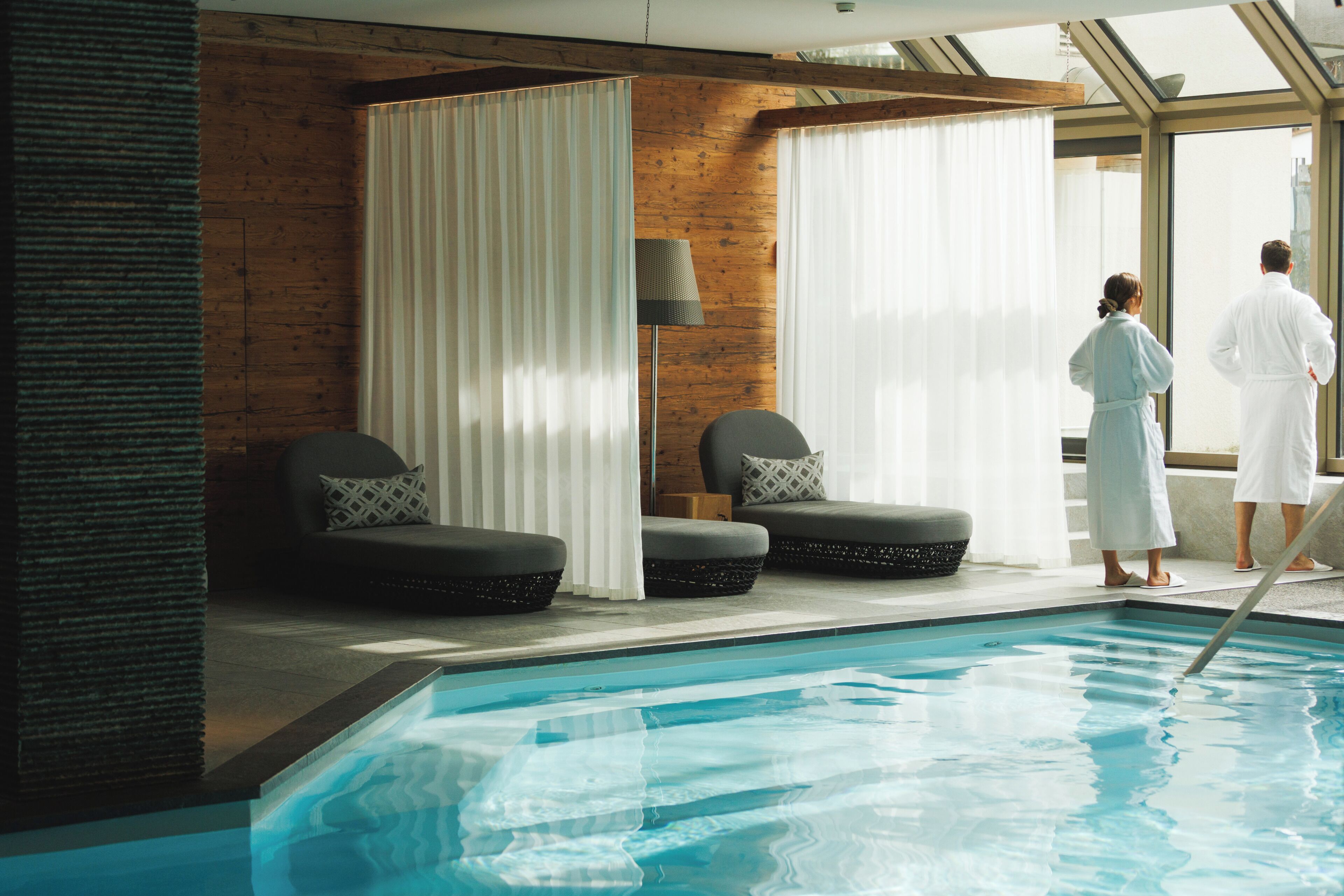 A swimming pool with two lounge chairs, a wooden wall, and a large window.