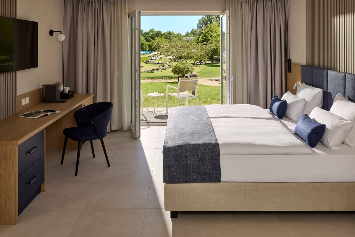 A modern hotel room with a large bed, a desk with a chair, a television, and a view of a garden and outdoor seating area.