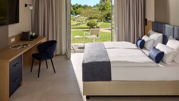 A modern hotel room with a large bed, a desk with a chair, a television, and a view of a garden and outdoor seating area.