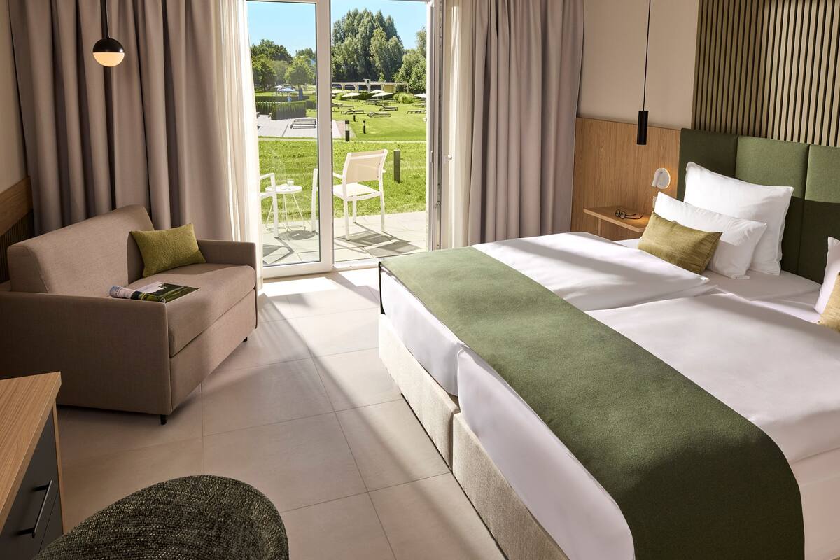 A hotel room with a sofa, a bed with a green blanket, a small table, and a view of a garden through a sliding door.