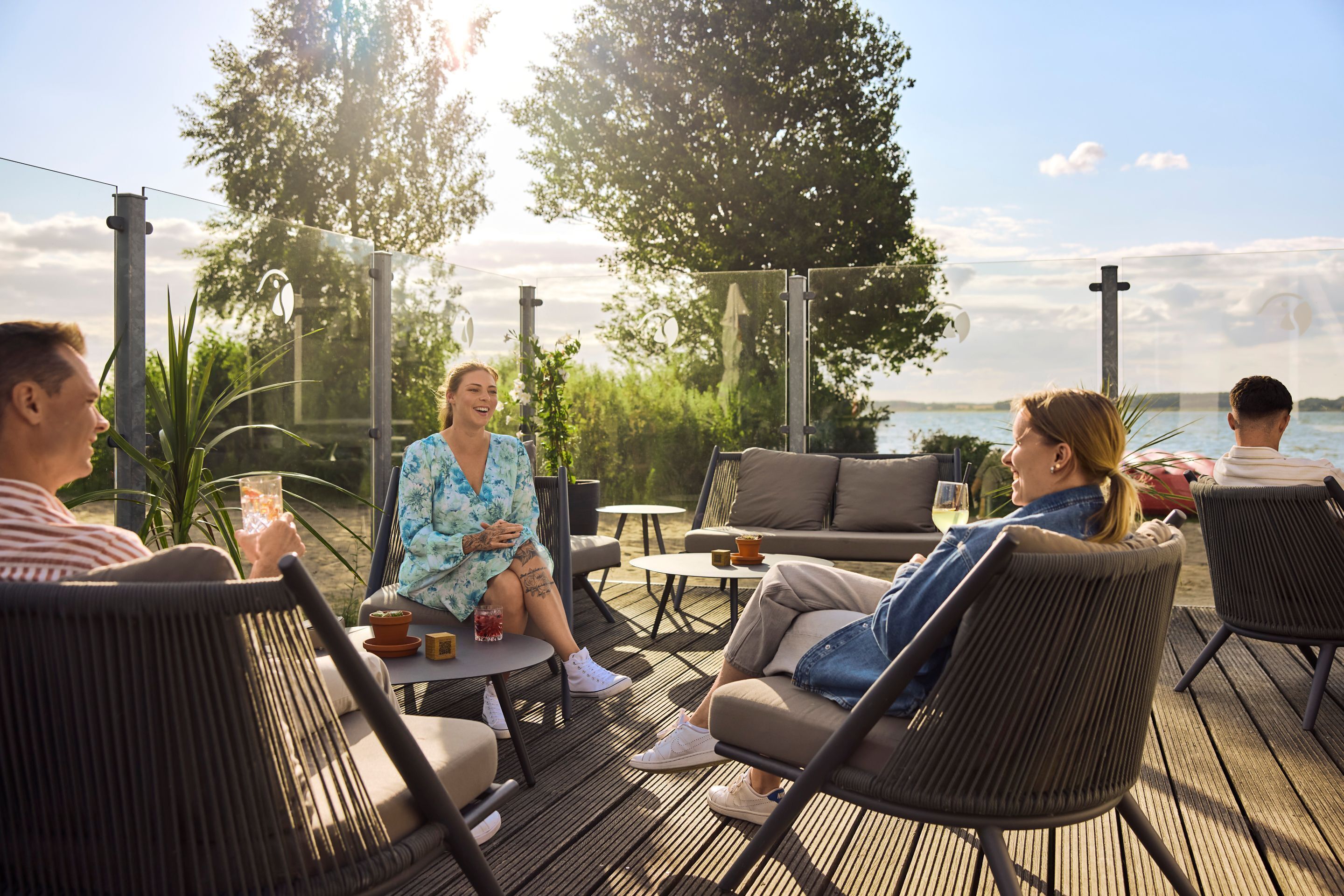 Un grupo de personas sentadas en muebles de exterior en una terraza de madera con vistas al mar.