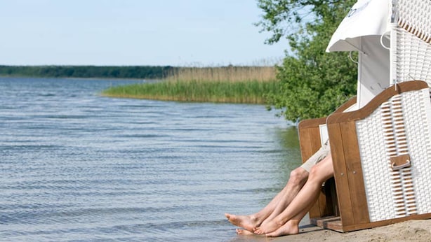 Une chaise de plage en bois recouverte d’une toile blanche, placée sur une plage de sable près d’une étendue d’eau.