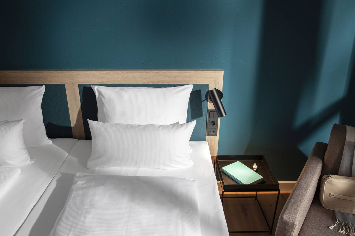 A hotel room with a wooden headboard, white bedding, a small bedside table with a lamp, and a chair.