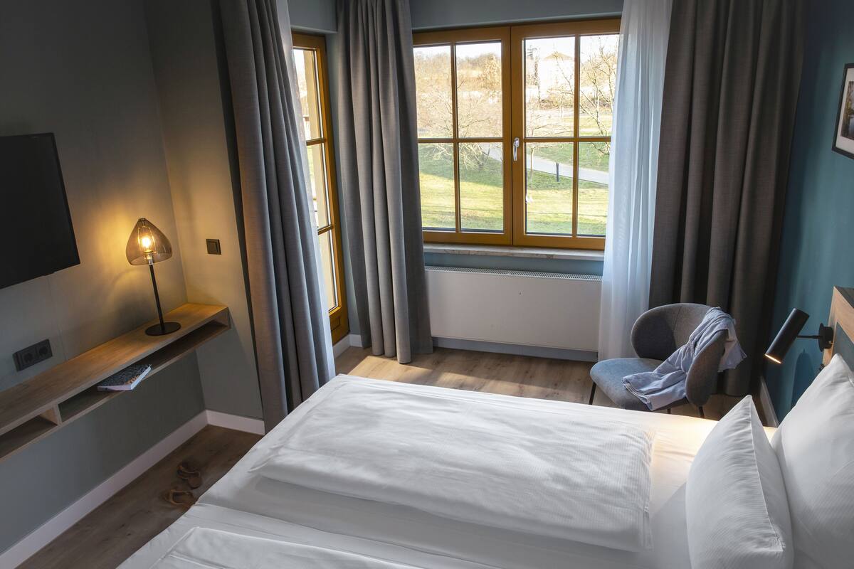 A bedroom with a bed, a chair, a TV, a window with curtains, and a lamp.