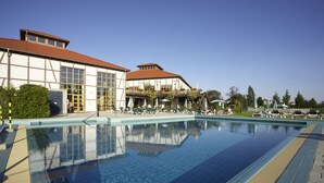 Une piscine avec des chaises longues et des parasols, un bâtiment doté de grandes fenêtres et d’un toit rouge, et un ciel bleu azur.