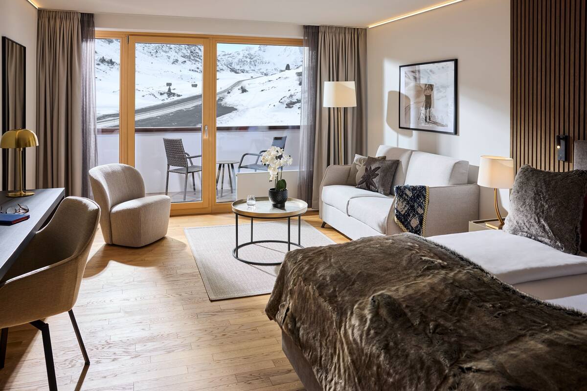 A modern hotel room with a large window, a sofa, a bed, a desk, and a chair.