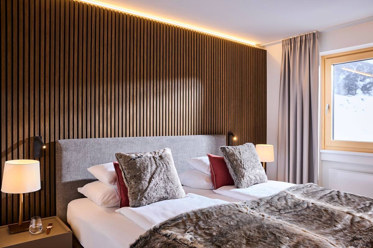 A hotel room with a large bed, two bedside lamps, a wooden panel wall, and a window with curtains.