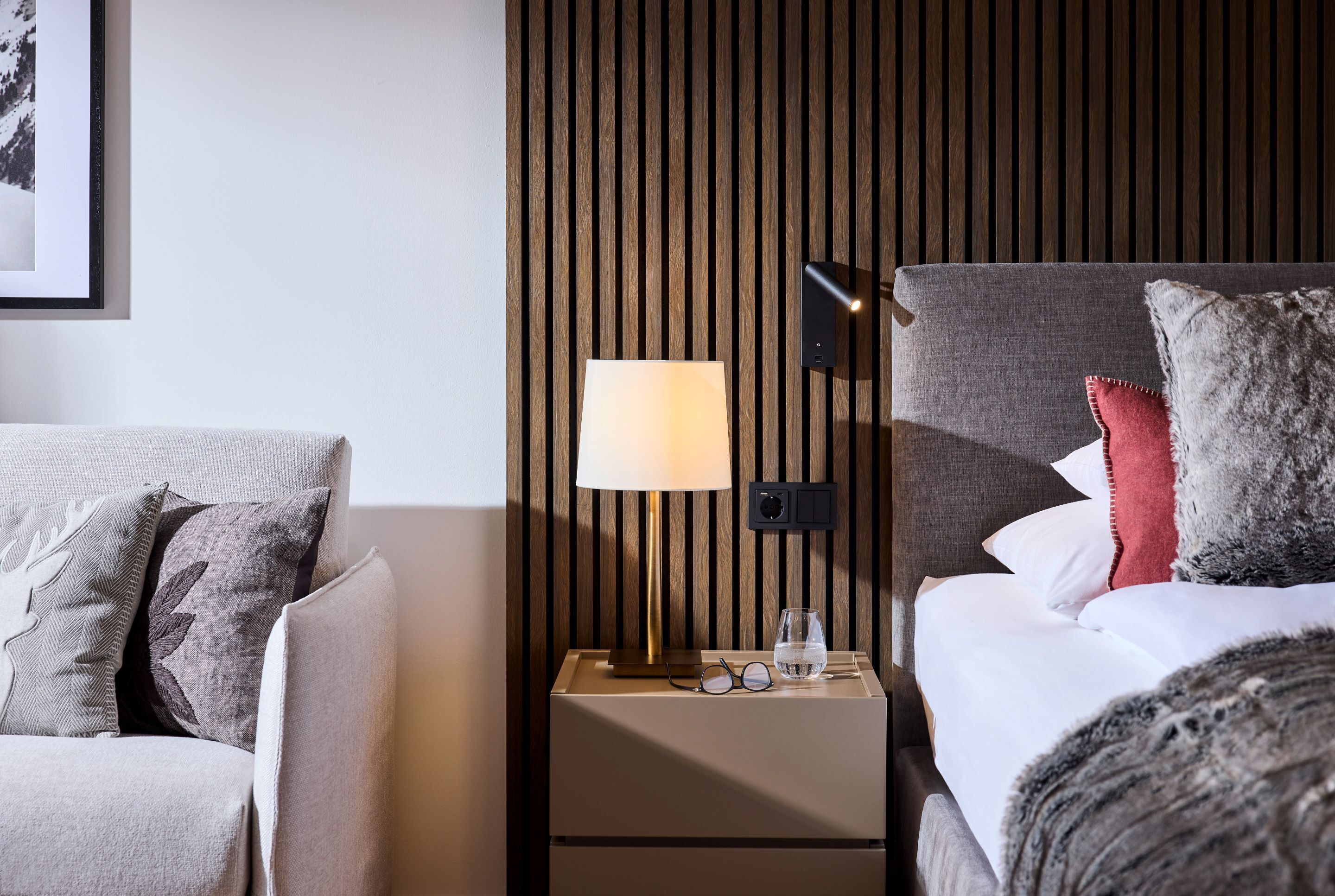 A modern hotel room with a wooden panel wall, a bedside table with a lamp, a glass of water, and a bed with pillows.