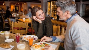 A couple dining at a restaurant with wooden tables and chairs, a bar in the background, and various dishes and drinks on the table. - ROBINSON ALPENROSE ZÜRS (Lech am Arlberg)