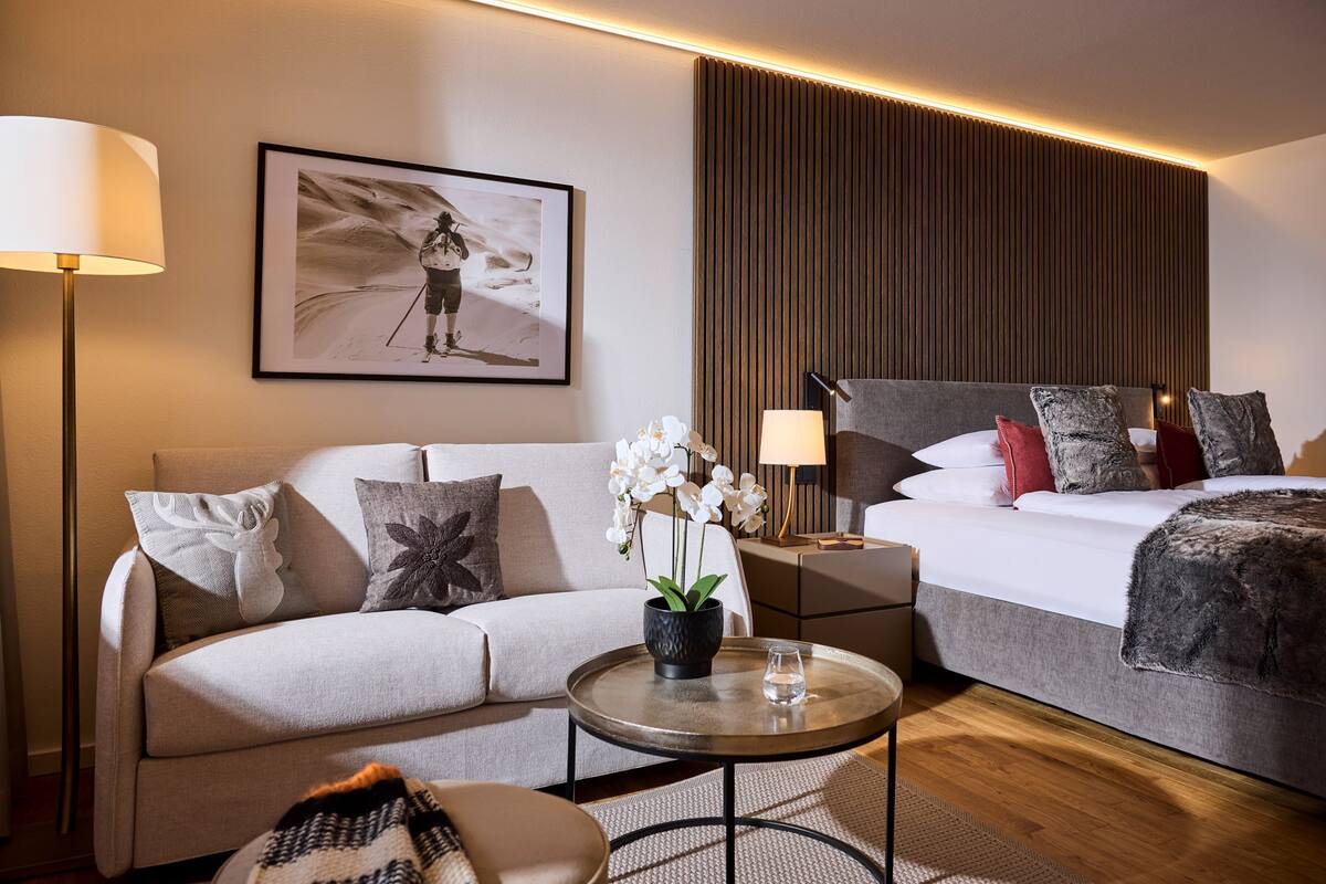 A modern hotel room with a sofa, a bed, a small table with a vase of flowers, and a framed picture on the wall.