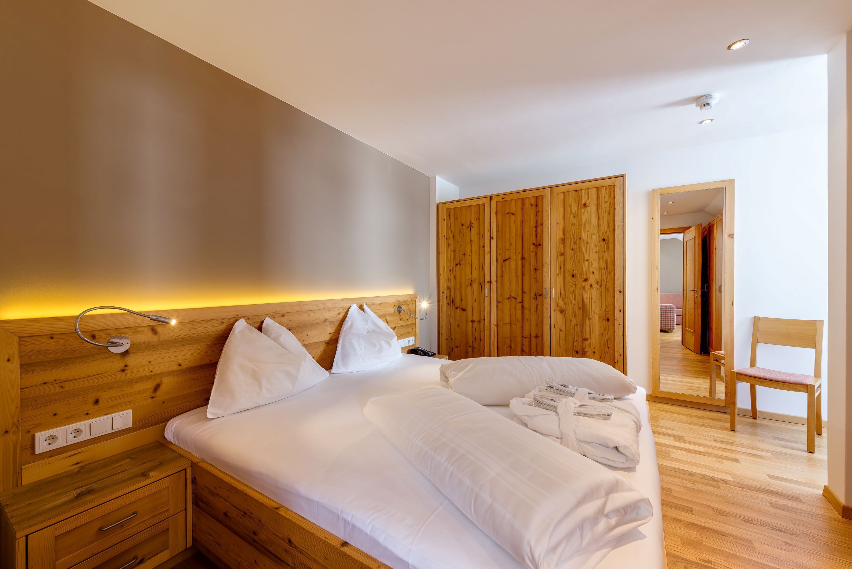 A bedroom with a wooden headboard, a large bed with white linens, a wooden wardrobe, and a mirror.
