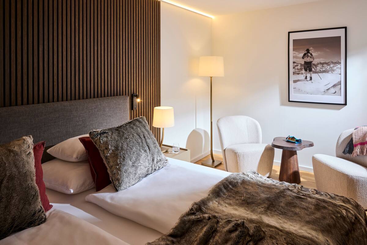 A modern hotel room with a large bed, a wooden headboard, a white armchair, a small round table, and a framed picture on the wall.