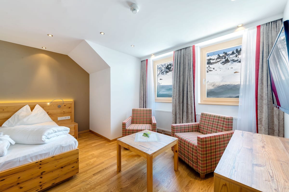 A hotel room with a wooden bed, two plaid armchairs, a small table, and a large window with a mountain view.