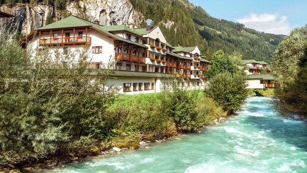 Un hôtel doté de nombreux balcons donnant sur une rivière, entouré d’une végétation luxuriante et de montagnes.