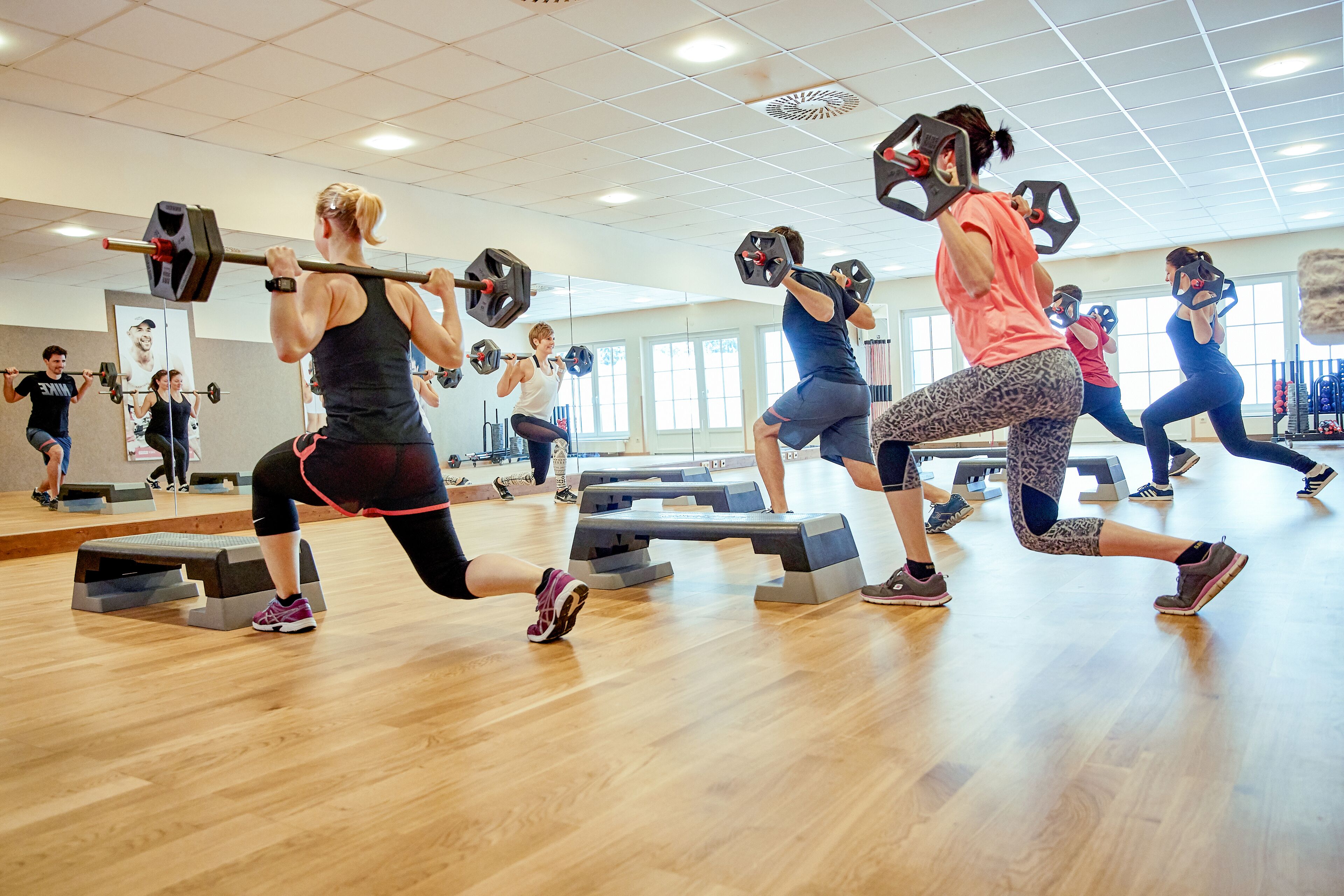 Eine Gruppe von Menschen trainiert mit Gewichten in einem Fitnessstudio mit Holzböden und Spiegeln.
