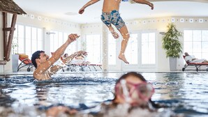 Een binnenzwembad met helder water, een duikplank en enkele mensen die zwemmen en springen.