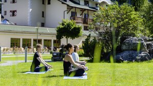 Un groupe de personnes assises sur des tapis de yoga dans un espace verdoyant, avec un bâtiment en arrière-plan.
