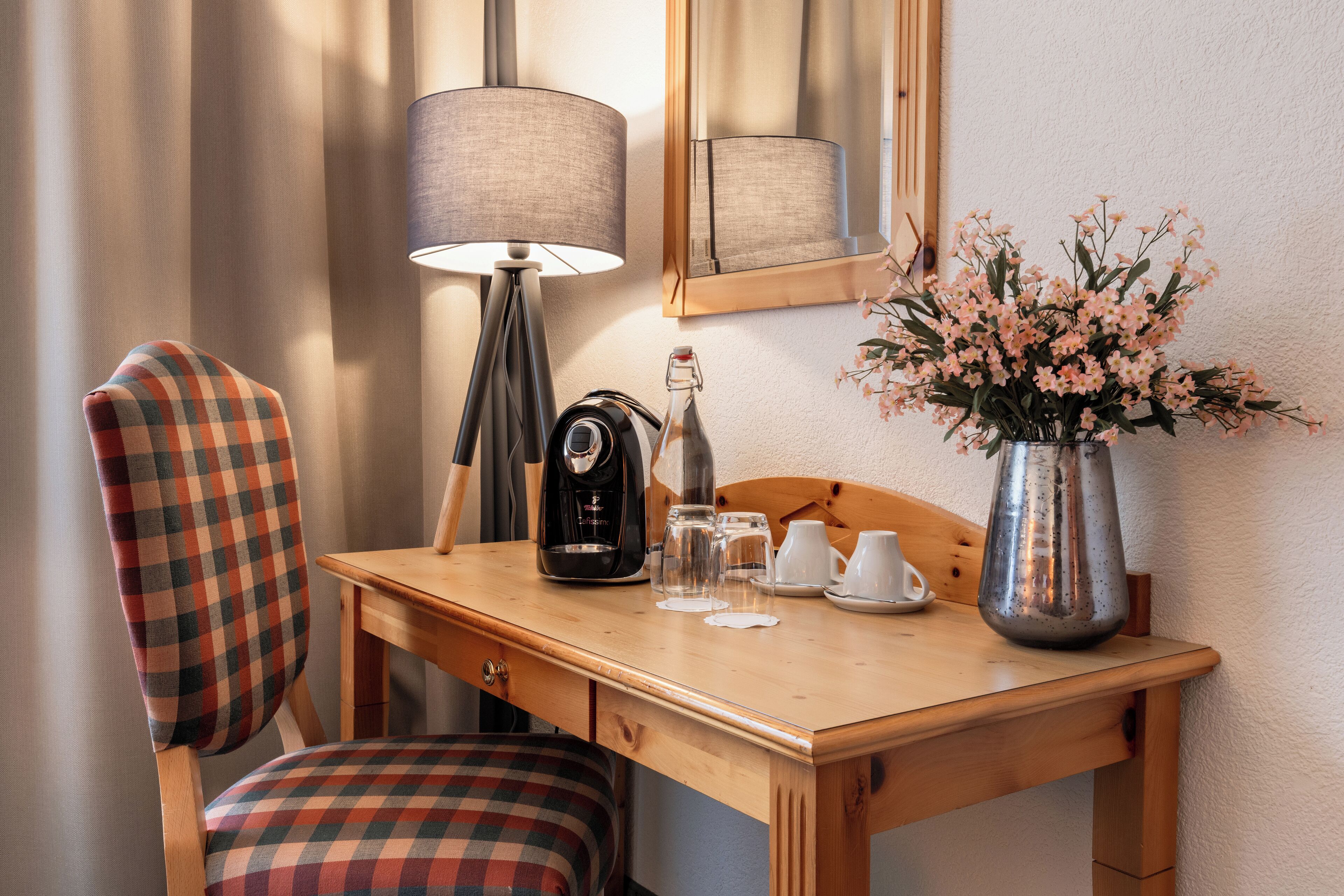 A wooden desk with a lamp, a coffee maker, a vase with flowers, and a chair with a plaid cushion.