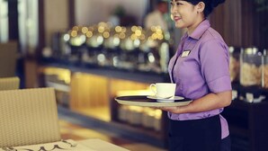 Une personne vêtue d’un uniforme violet, tenant un plateau sur lequel se trouvent une tasse et une soucoupe, dans une pièce dont l’arrière-plan est flou et où l’on aperçoit un comptoir éclairé.