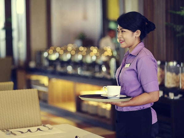 Una persona con uniforme morado sostiene una bandeja con una taza y un platito, en una habitación con un fondo desenfocado que muestra una barra con luces.
