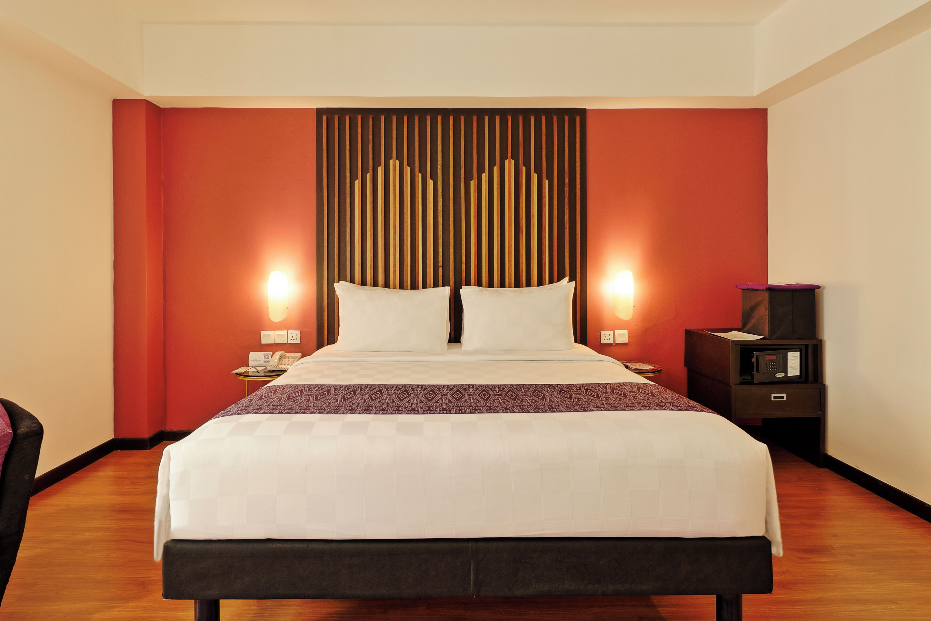 A hotel room with a large bed, two bedside lamps, a nightstand with a radio, and a red accent wall.