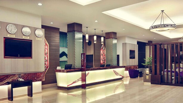 Un hall d’hôtel moderne, avec un comptoir d’accueil, des horloges indiquant l’heure de Tokyo, de Londres et de New York, et un grand lustre.
