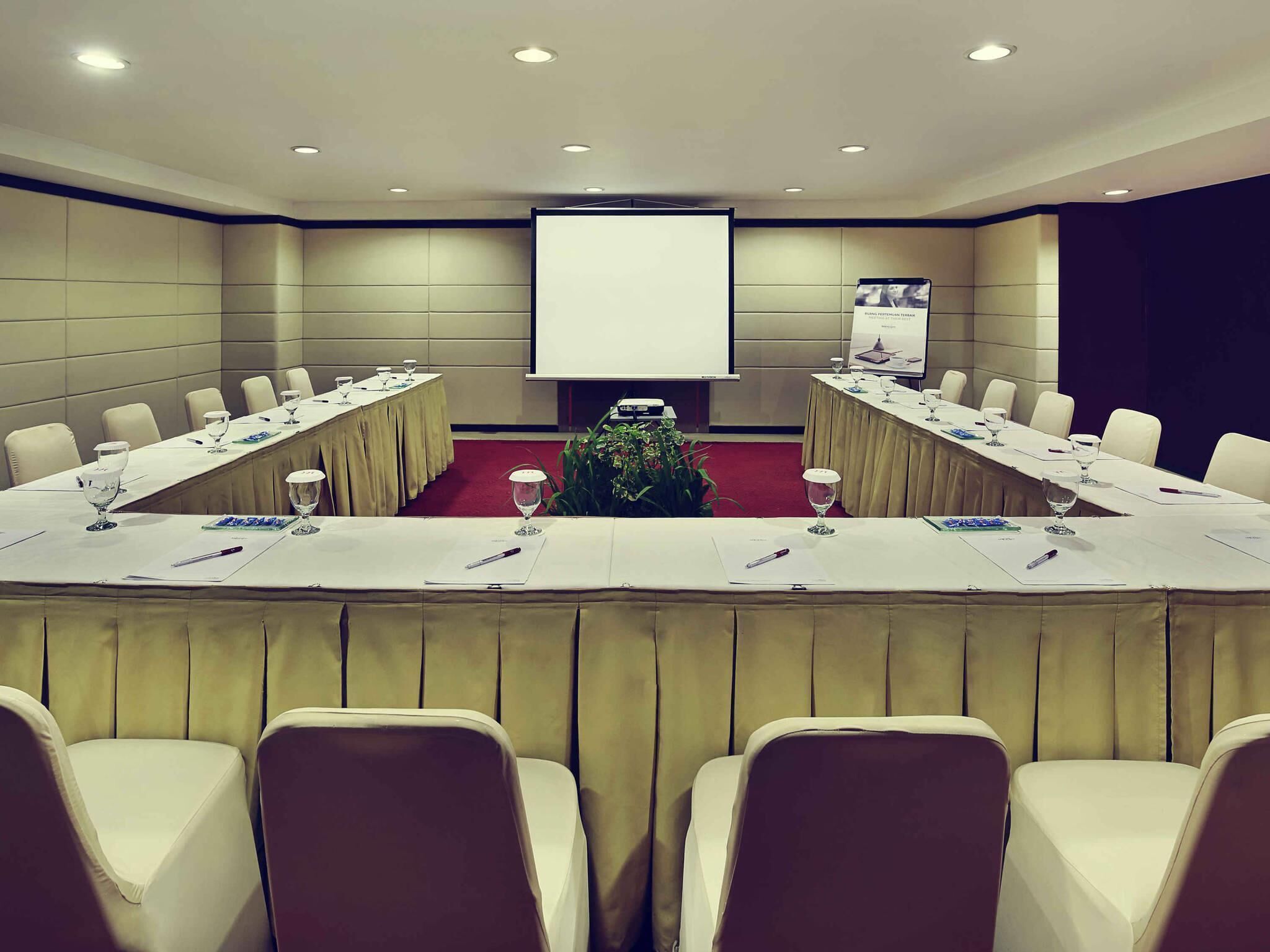 Un salón de conferencias con una larga mesa, sillas, una pantalla de proyector y un arreglo floral.