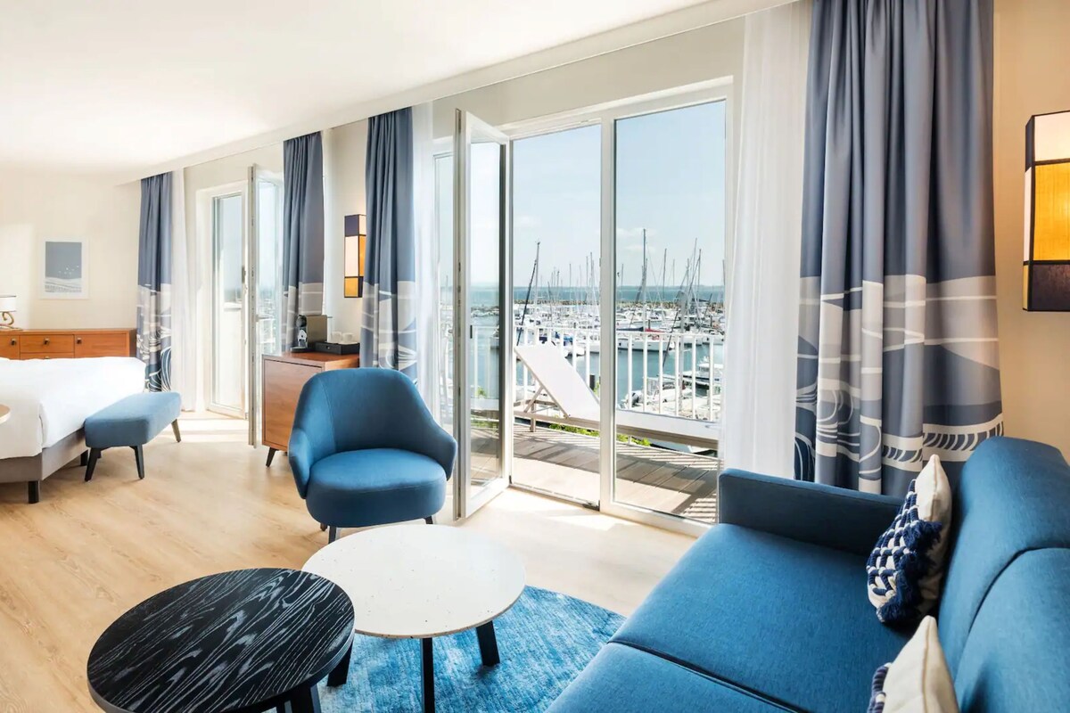 A hotel room with a bed, a desk, a chair, a sofa, and a view of a marina.