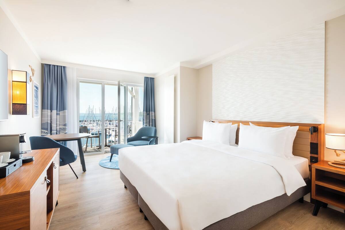 A hotel room with a large bed, a desk with a computer, a chair, and a view of the city through large windows.