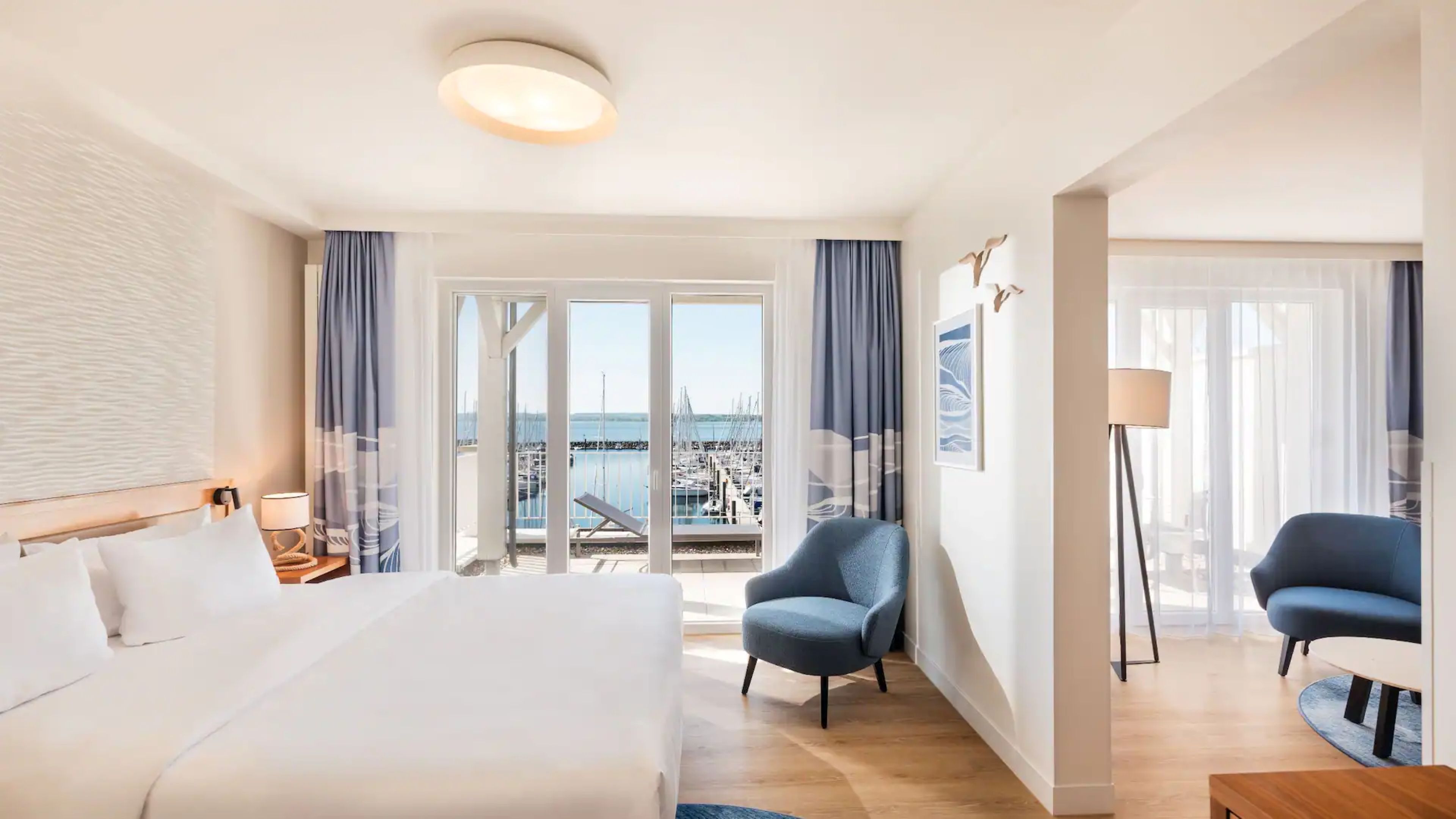 Habitación de hotel con cama, dos sillones, un escritorio, una lámpara y vista al puerto deportivo.