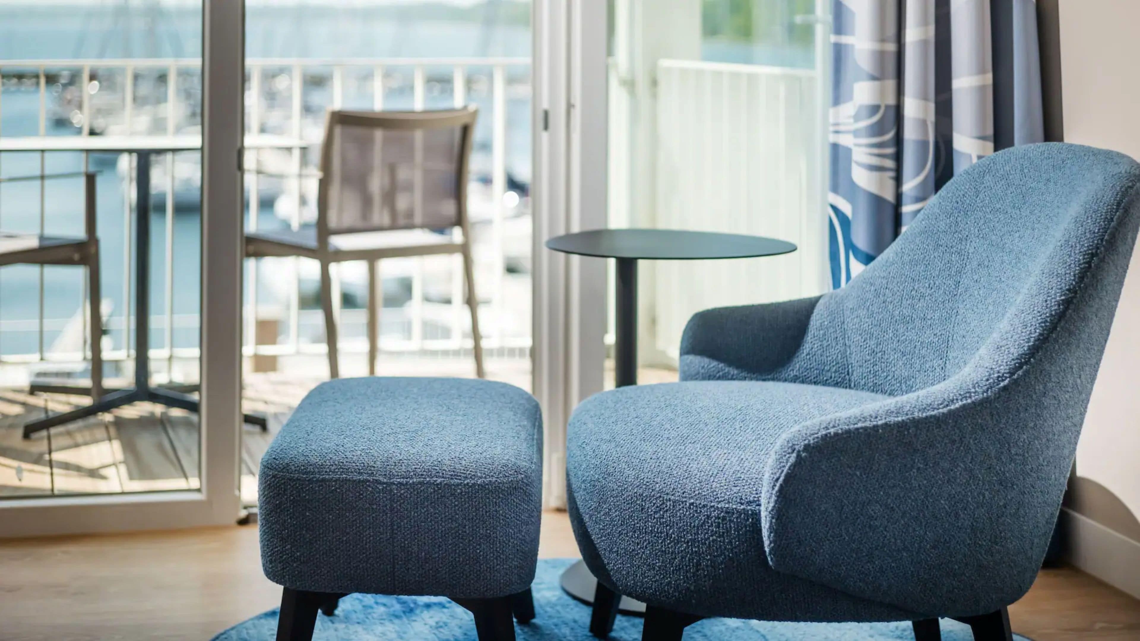 A room with a blue armchair and ottoman, a small round table, and a view of the sea through a glass door.