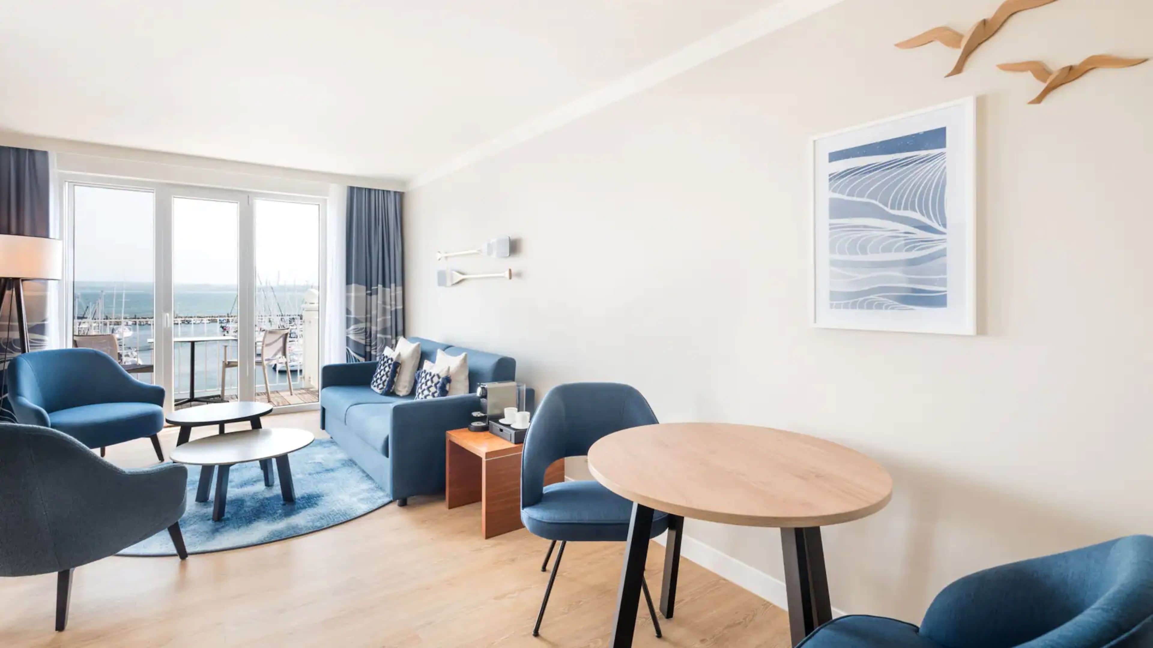 Ein modernes Wohnzimmer mit einer Couch, Sesseln, einem Couchtisch und Blick auf den Yachthafen.