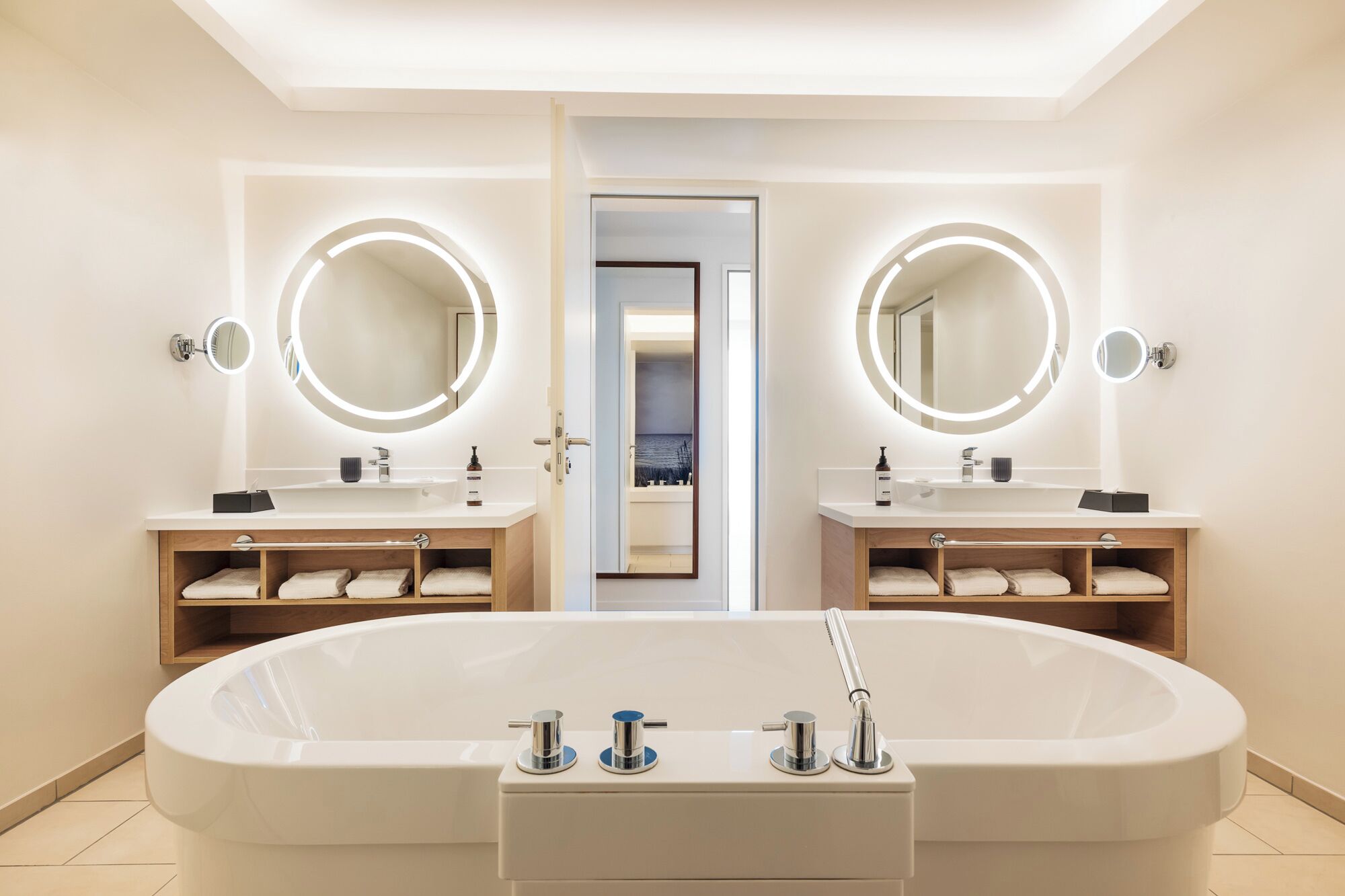 Une salle de bain moderne avec une grande baignoire sur pieds, deux vasques avec des miroirs ronds et des étagères intégrées où sont rangées les serviettes.