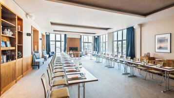 Ein Konferenzraum mit langen Tischen, Stühlen und einem großen Bildschirm. An einer Seite stehen Bücherregale und auf der anderen eine blickdichte Fensterschranke.