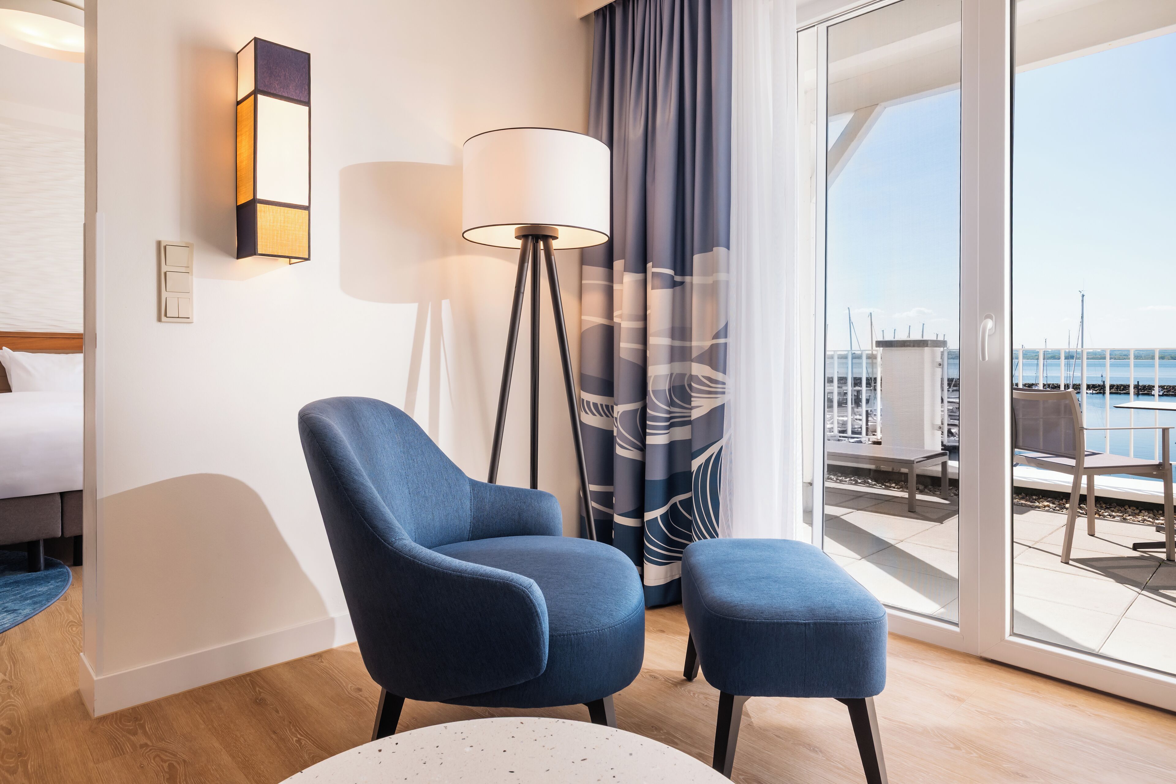 A modern hotel room with a large window, a blue armchair, a small ottoman, a floor lamp, and a bed with a white headboard.
