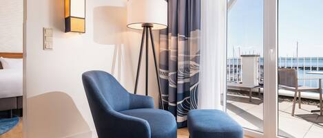 Moderni hotellihuone, jossa on suuri ikkuna, sininen nojatuoli, pieni jalkarahi, lattiavalaisin ja valkoinen päätyosalla varustettu sänky.