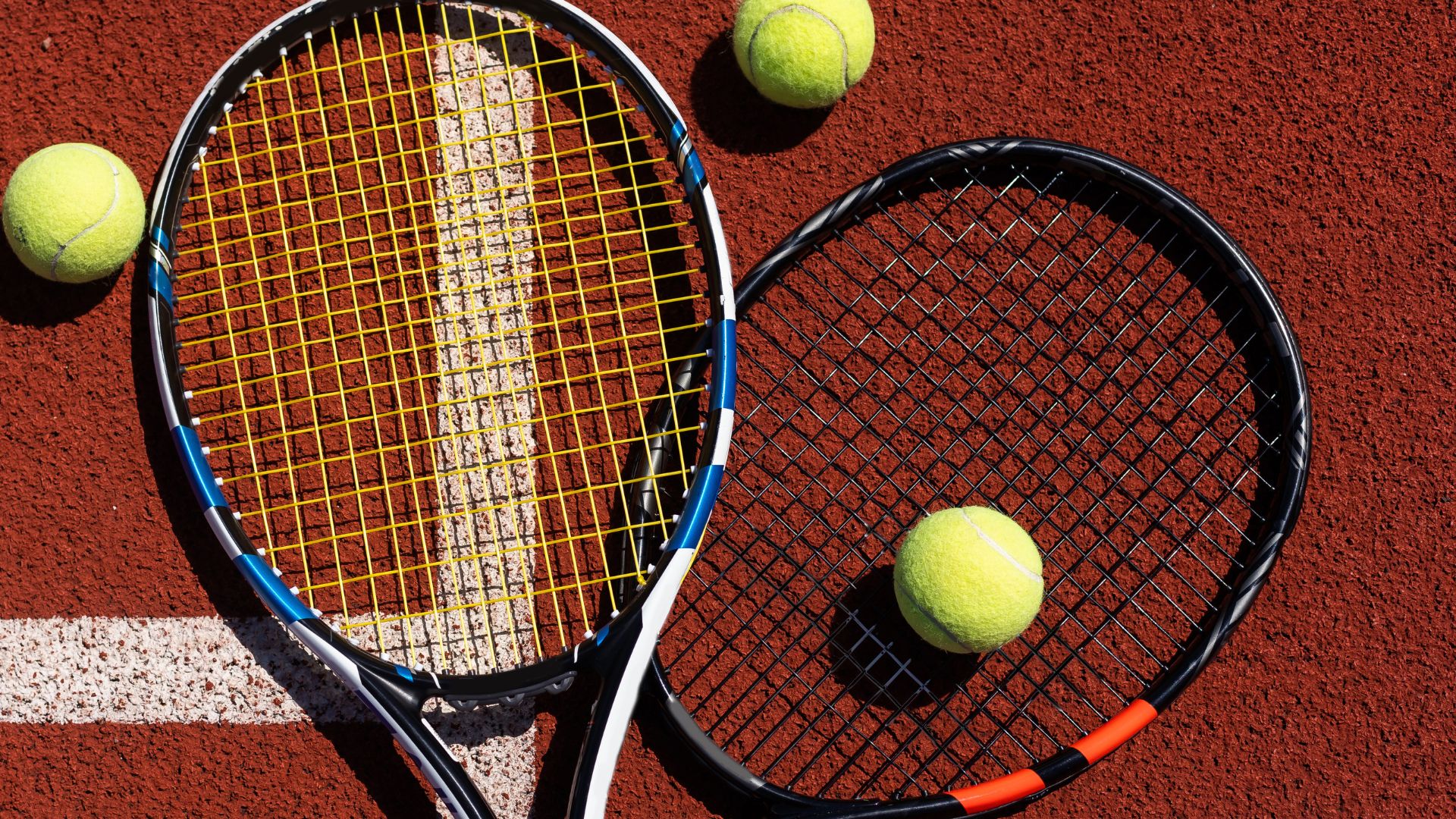 Two tennis rackets and three tennis balls on a red clay court.