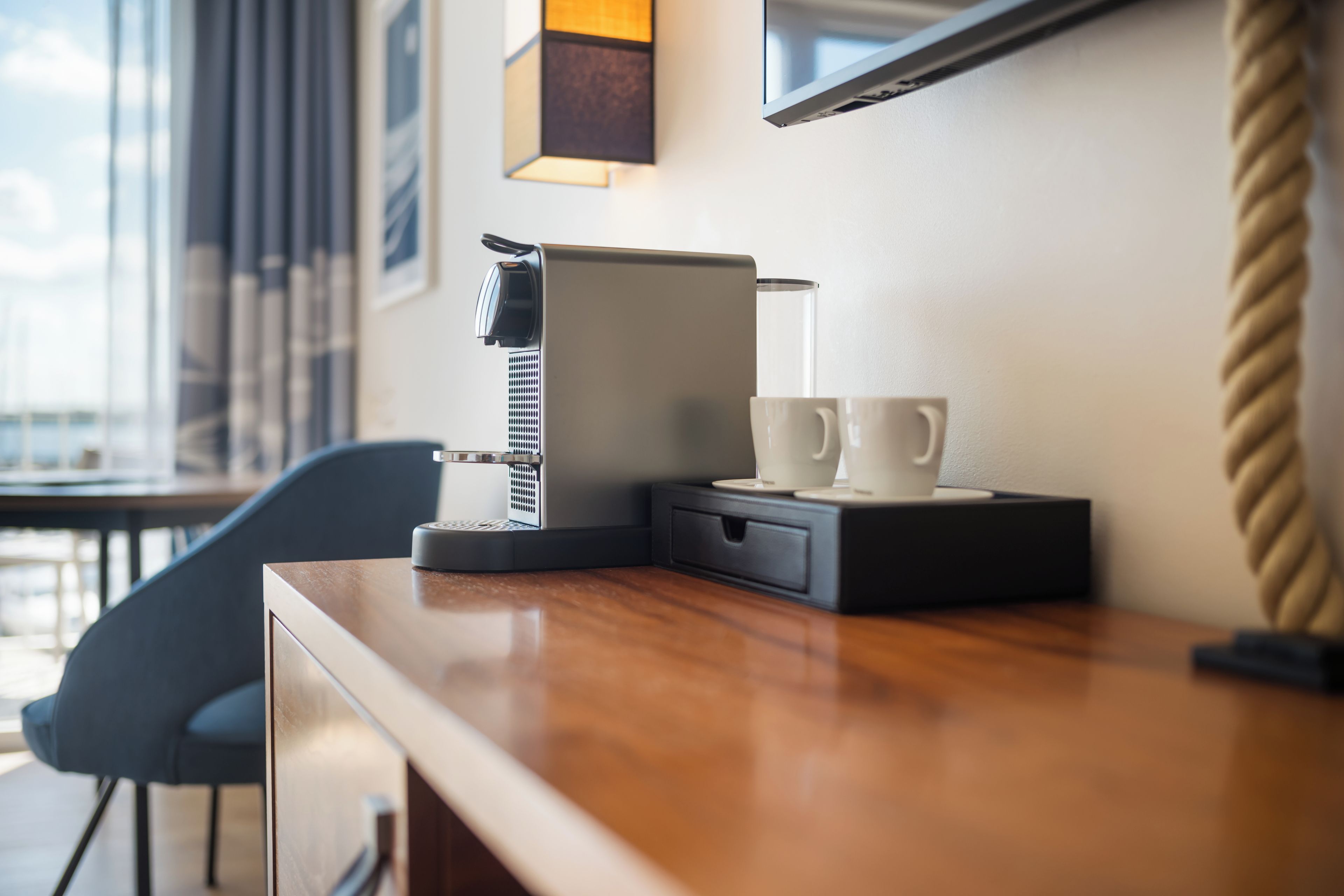 Una cafetera con un recipiente de goteo, dos tazas blancas y un cajón pequeño sobre una encimera de madera.