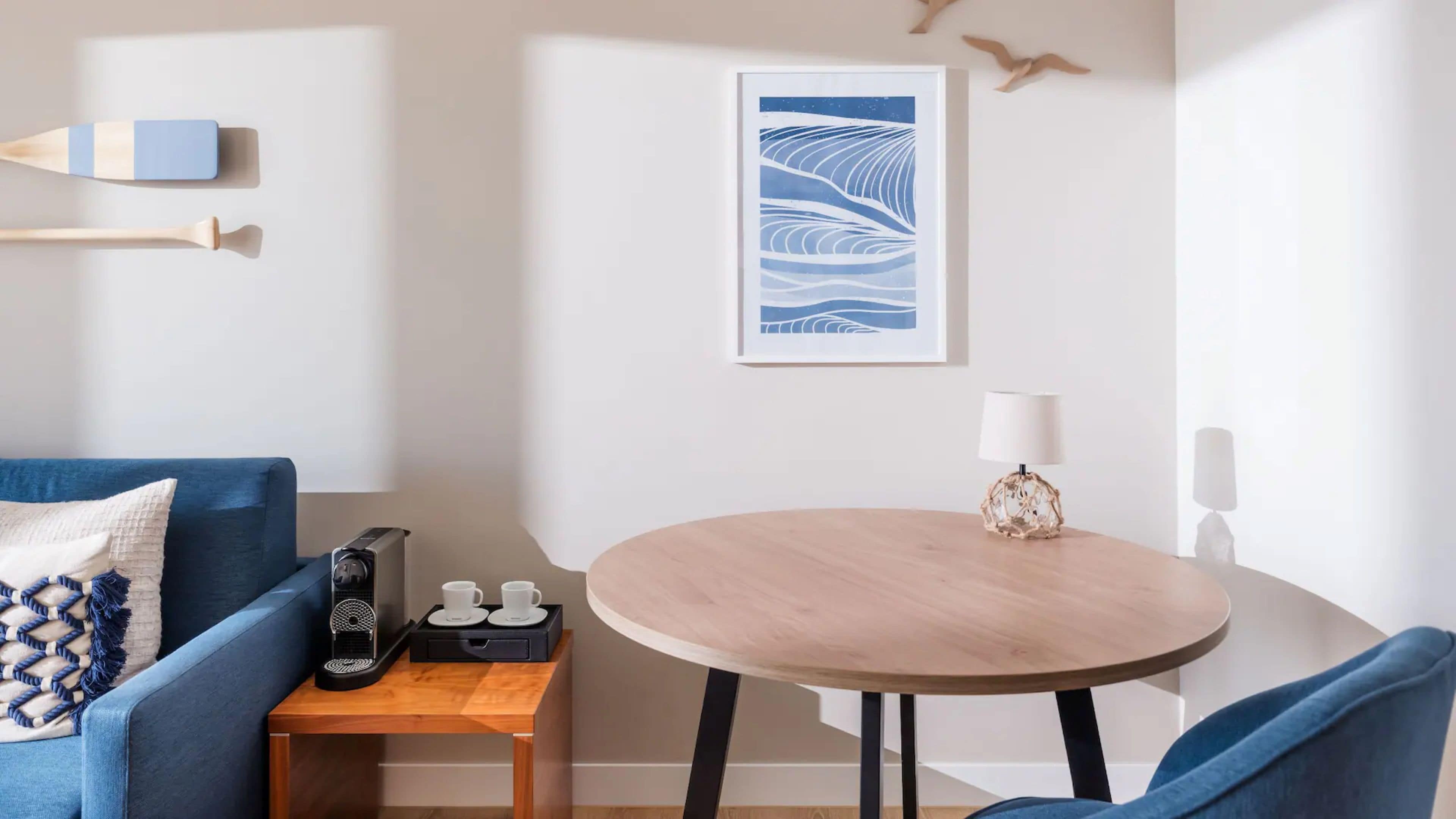 Ein modernes Zimmer mit einem blauen Sofa, einem kleinen Holztisch mit zwei Tassen, einer Kaffeemaschine und einem runden Tisch mit einer Lampe.