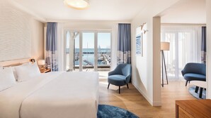 A hotel room with a large bed, two armchairs, a floor lamp, and a view of the marina.