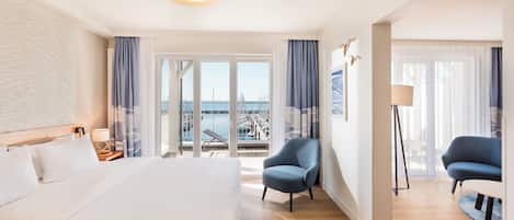 Habitación de hotel con una cama grande, dos sillones, una lámpara de pie y vistas al puerto deportivo.