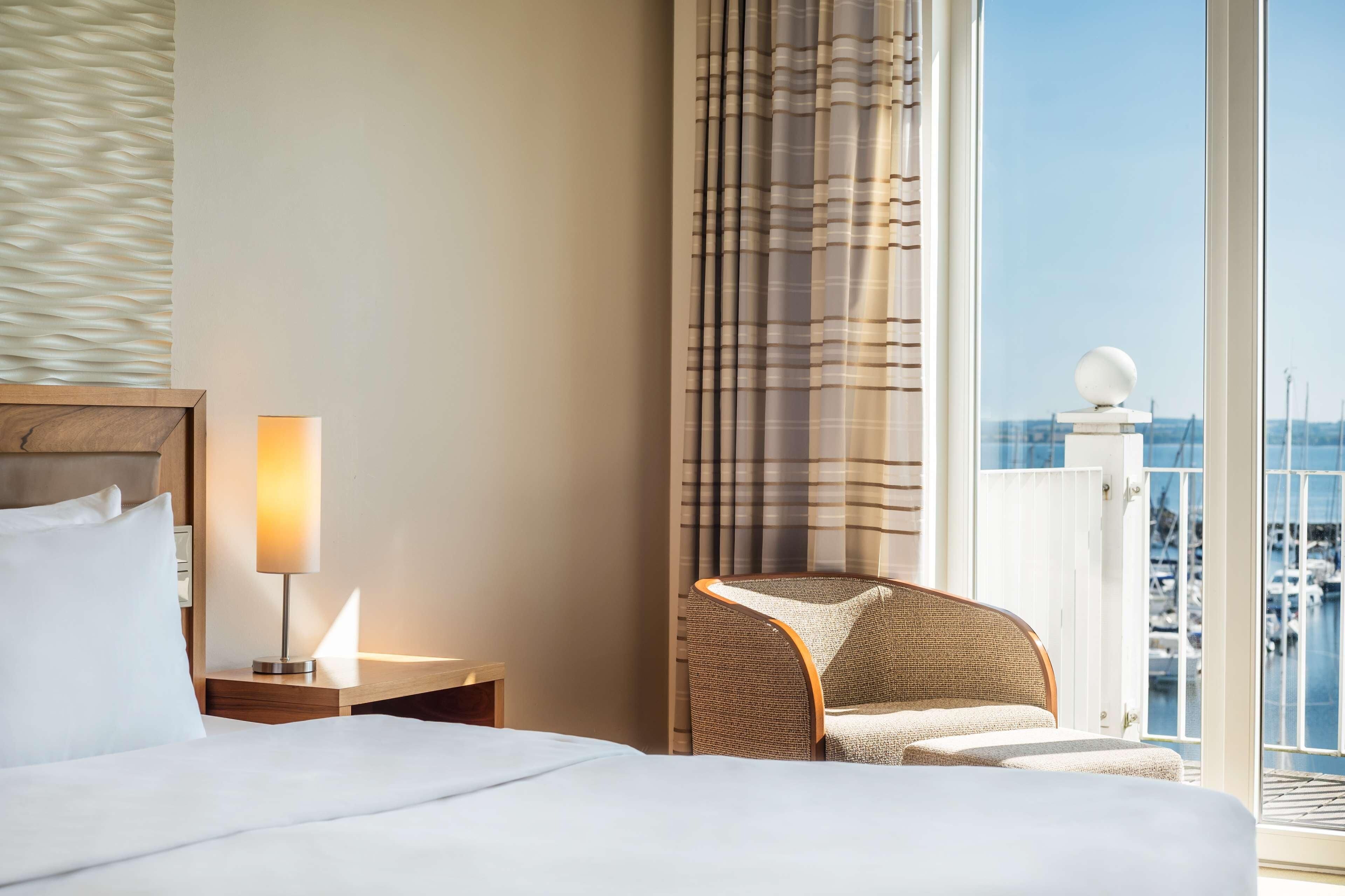 A hotel room with a bed, a nightstand with a lamp, a chair, and a view of a marina through a window.