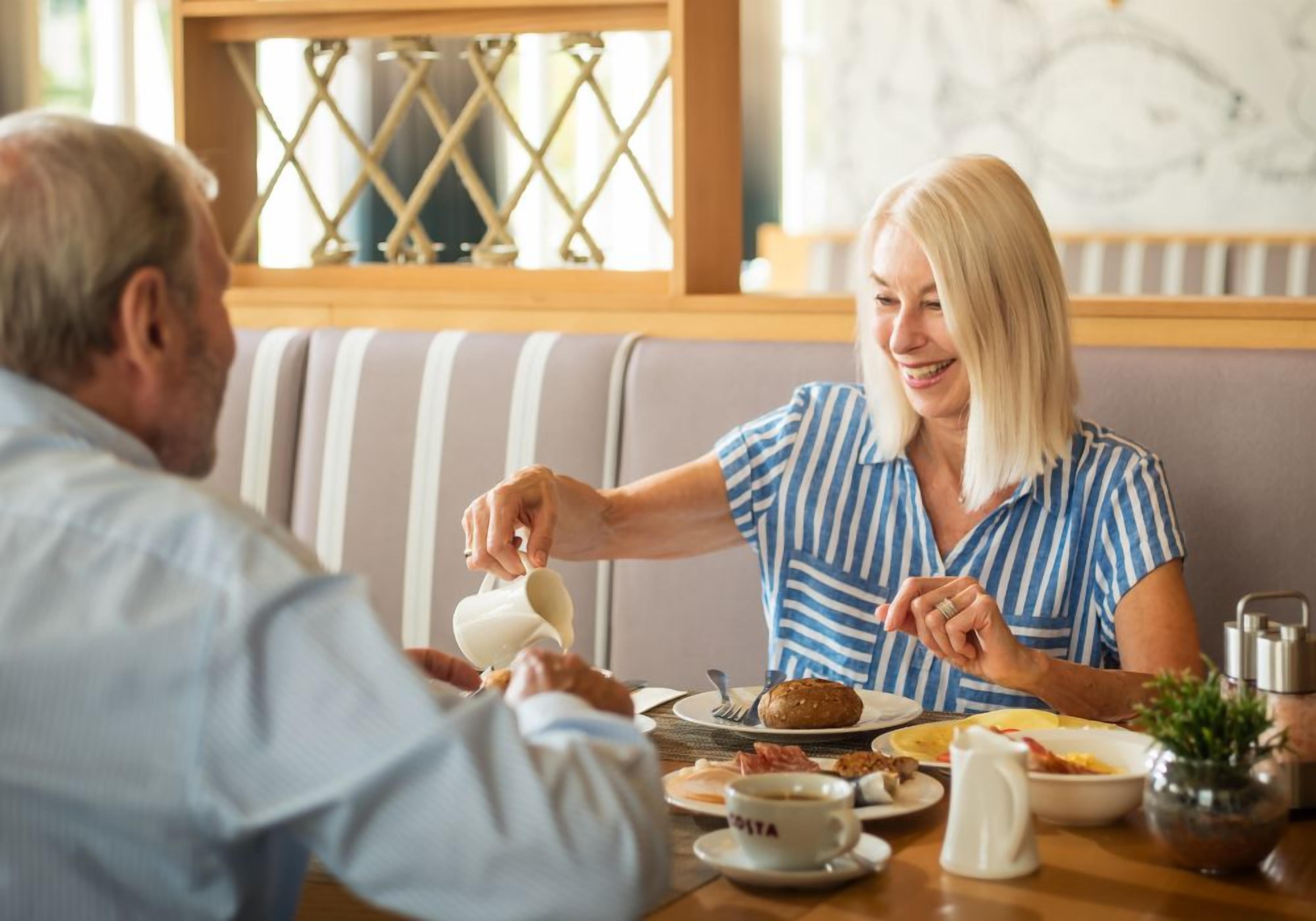Un coin repas avec une table dressée pour deux personnes, comprenant des assiettes avec de la nourriture et des boissons, une tasse sur laquelle est inscrit le mot « DATA », et une fenêtre ornée d’un motif en croisillons.
