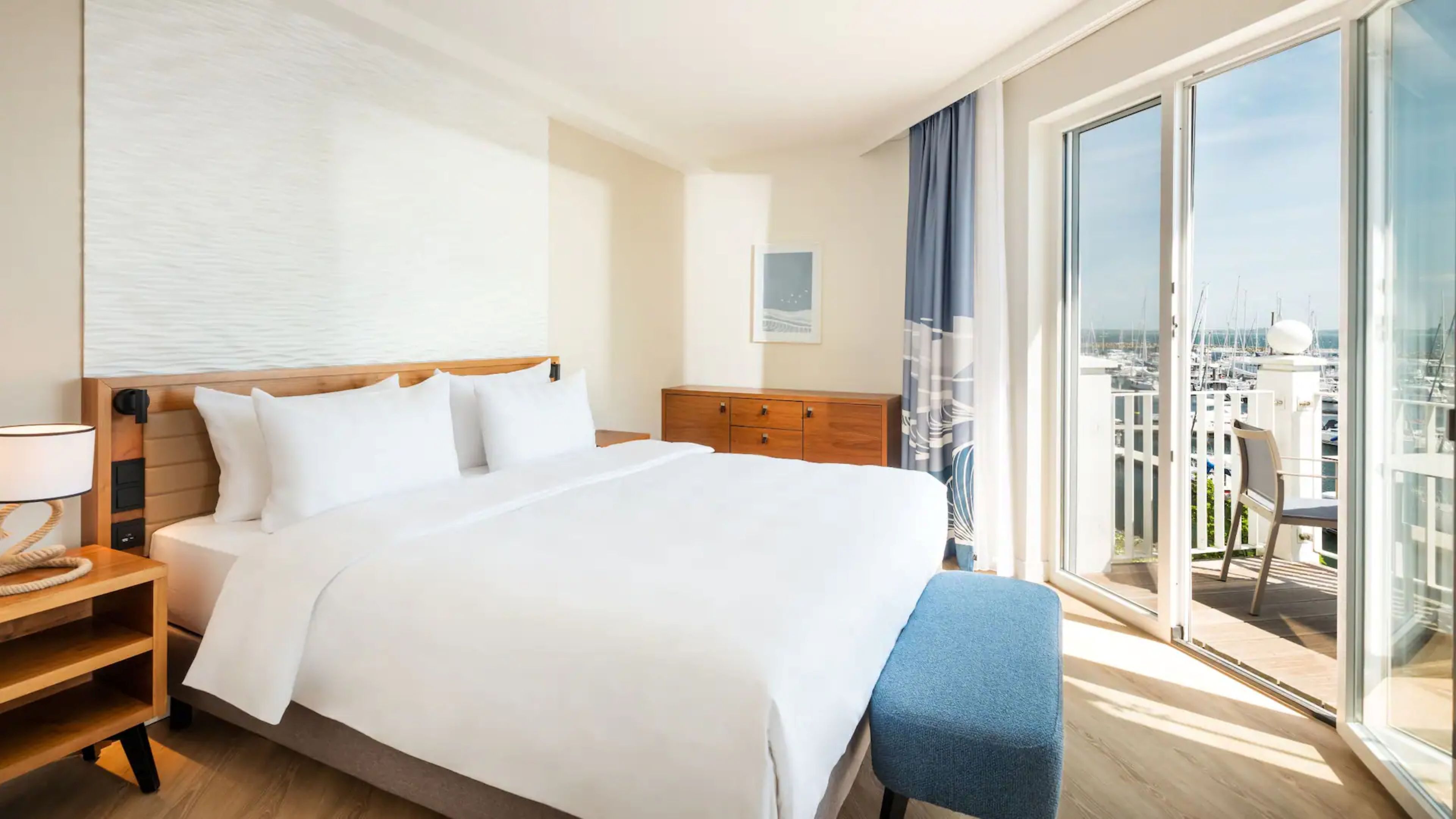 A hotel room with a large bed, a wooden nightstand, a dresser, and a view of a marina through a large window.